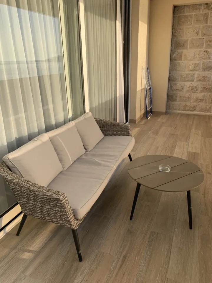 Ein gemütlicher Außenbalkon mit Rattansofa, Kissen und einem kleinen Tisch auf Holzboden.