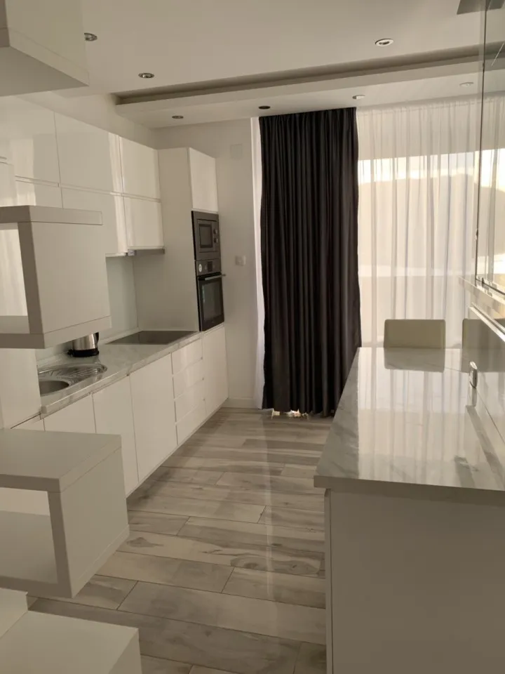 Moderne weiße Küche mit Hochglanzschränken, Esstisch und großem Fenster mit Vorhängen.
