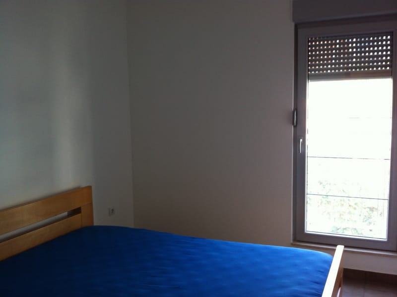 Schlafzimmer in einer Wohnung in Petrovac, Montenegro: Holzbett mit blauer Tagesdecke und Zugang zum Balkon mit Rollläden.