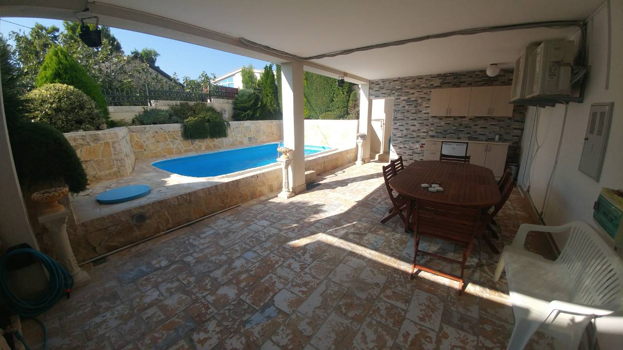 Patio couvert avec table à manger, chaises, cuisine extérieure et vue sur la piscine dans le jardin.