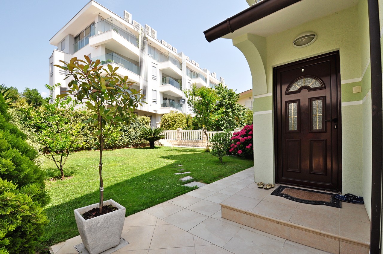 Eingang zum Haus mit grünem Garten und Terrasse in Tivat, Montenegro. Im Hintergrund ein moderner Wohnkomplex mit Balkonen.