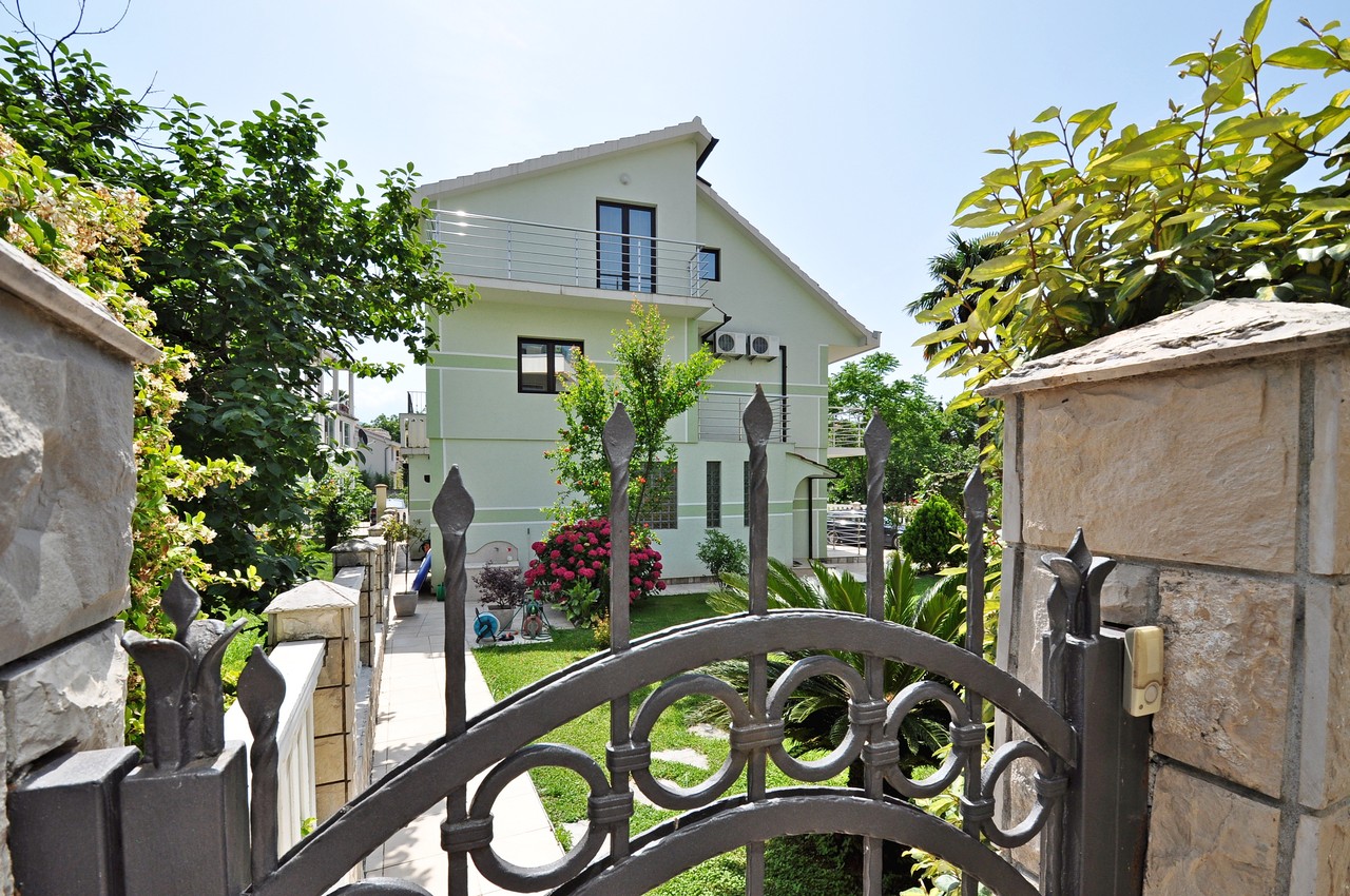 Dreistöckiges Haus mit Garten und schmiedeeisernem Tor in Tivat, Montenegro, Blick auf die Fassade mit Terrassen und grünem Grundstück.