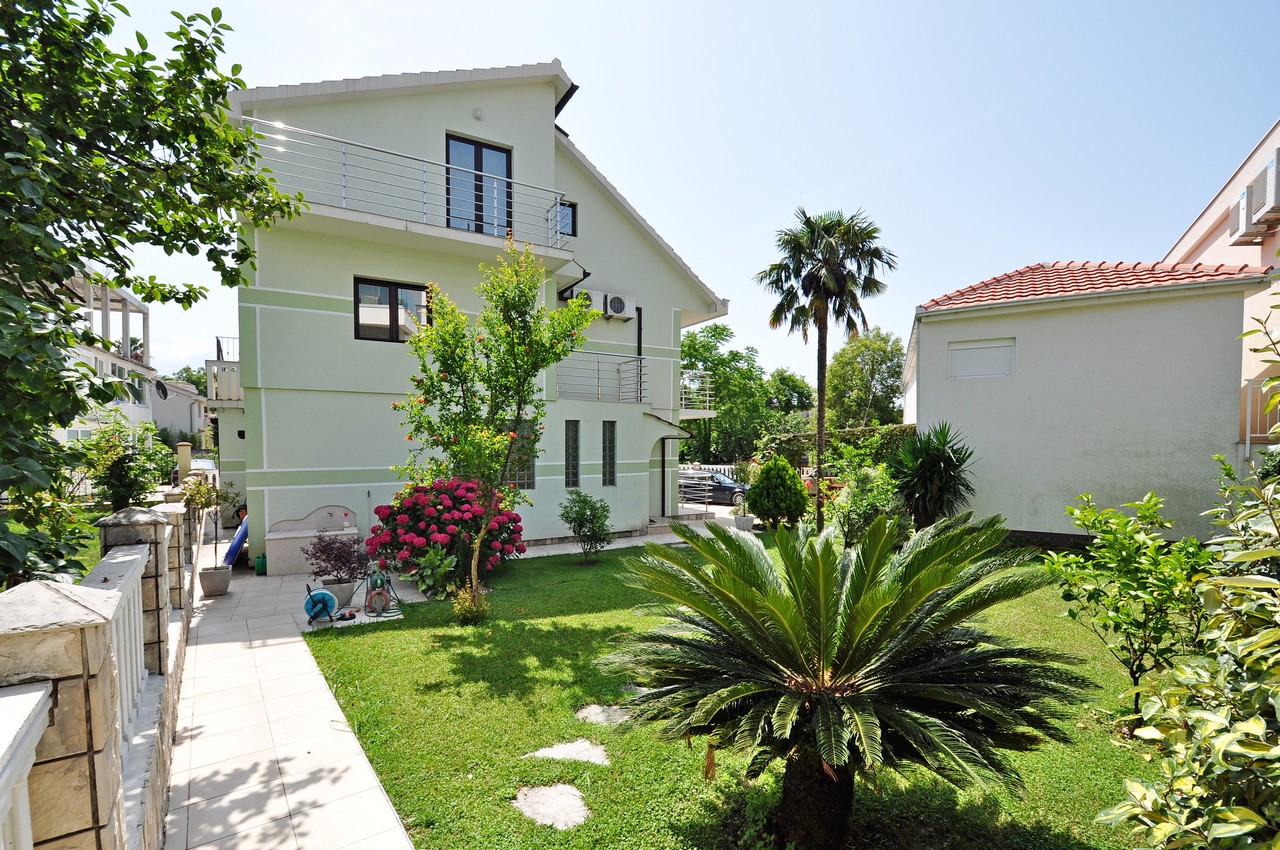 Dreistöckiges Haus in Tivat, Montenegro, mit gepflegtem Garten, Palmen, blühenden Sträuchern und Terrassen an der Fassade.