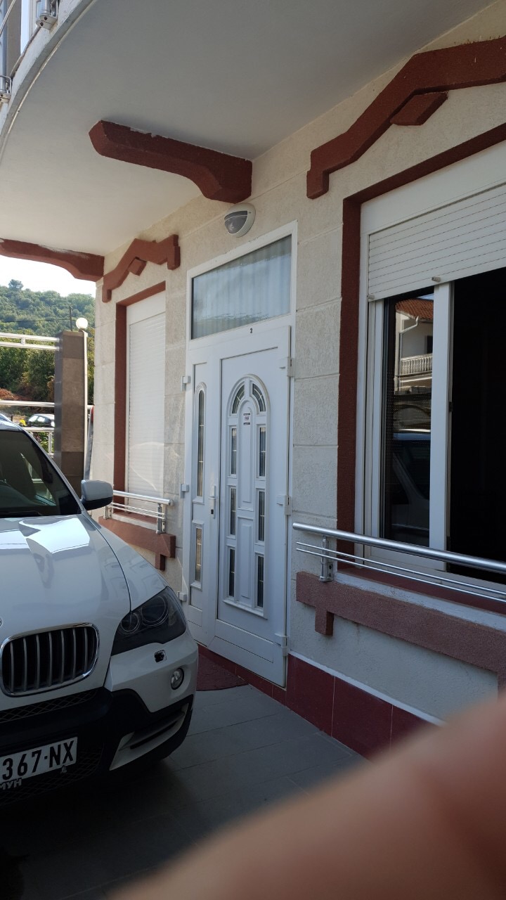 Ulaz u kuću u Tivtu, Crna Gora: bela plastična vrata, prozori sa roletnama i parking mesto ispred fasade.