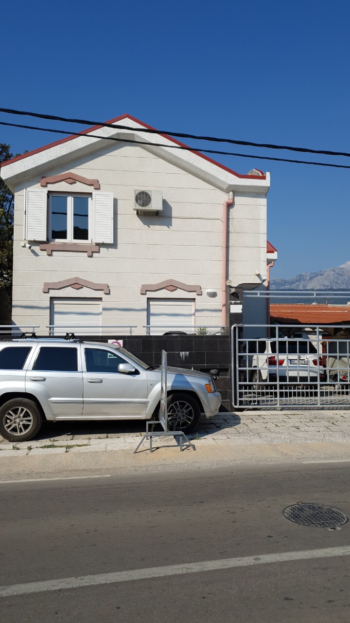 Dvospratna kuća u Tivtu, Crna Gora: fasada sa svetlom oblogom, metalna kapija i parking pored puta.