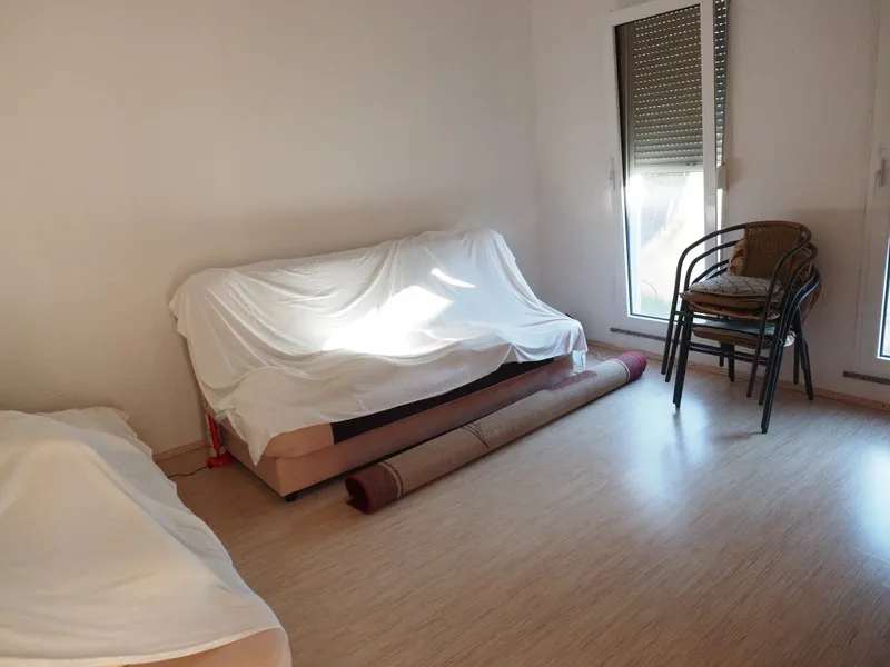 Svetla dnevna soba u kući u gradu Bar, Crna Gora: sofa s navlakom, laminat, stolice i smotan tepih pored prozora.