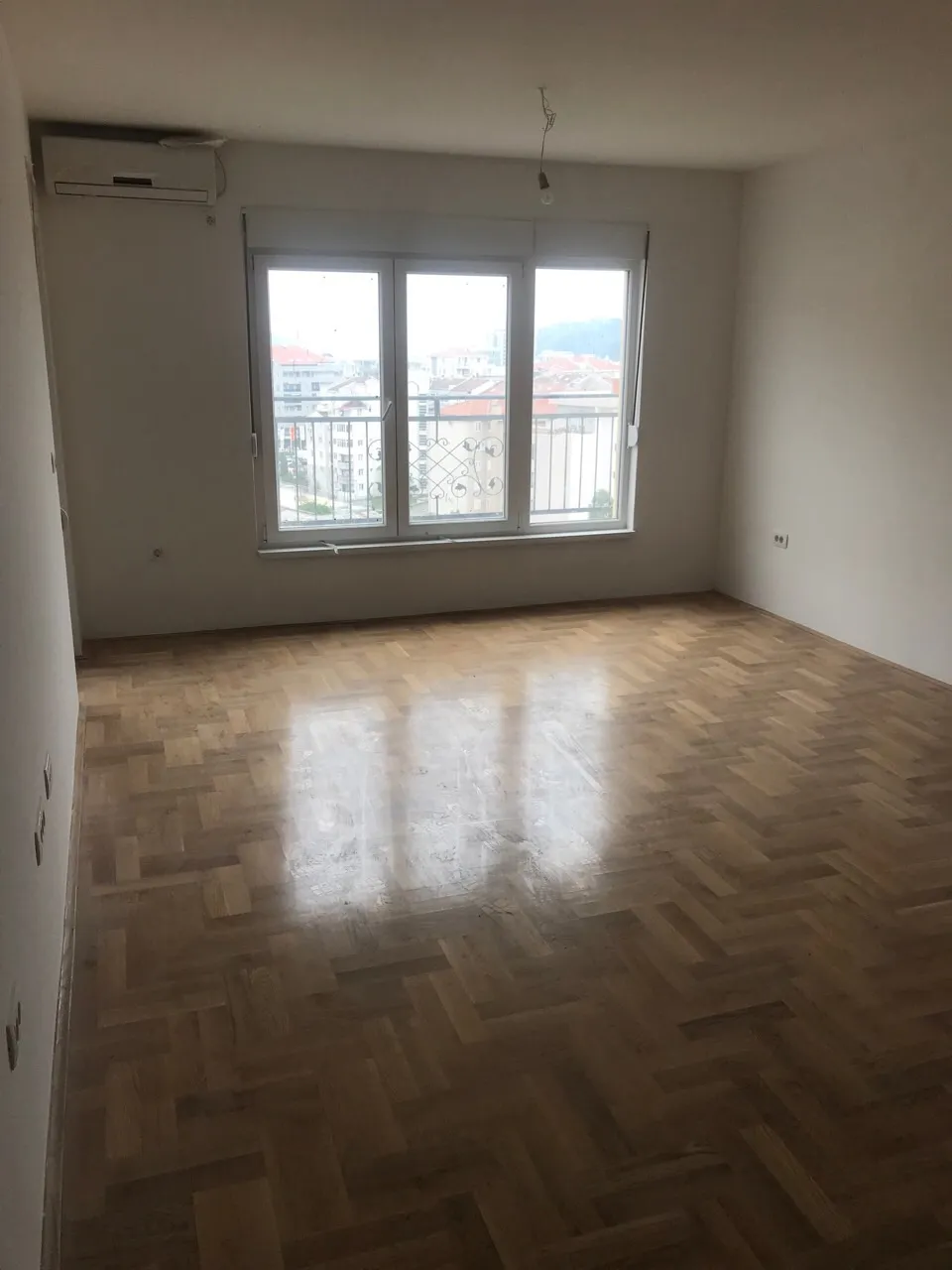 Helles Wohnzimmer in einer Wohnung in Budva, Montenegro: Parkettboden, weiße Wände, Klimaanlage und Stadtblick aus dem Fenster.