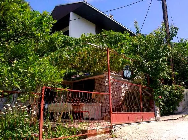 Dvospratna kuća u Šušnju, Crna Gora, sa terasom obraslom vinovom lozom, crvenom ogradom i zelenim vrtom na placu.