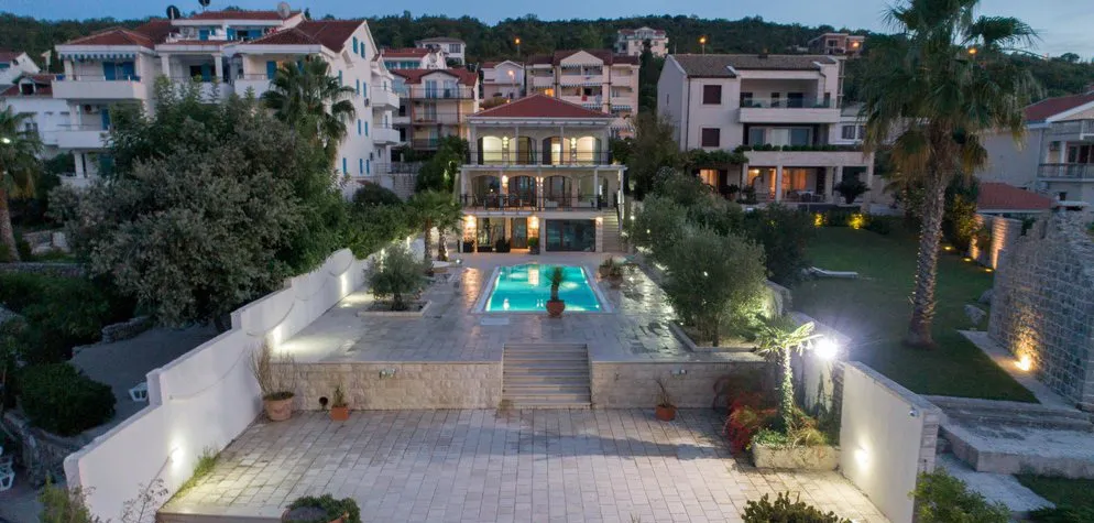 Villa with a pool and terrace in Krašići, Montenegro: view of the illuminated house facade and landscaped grounds in the evening.