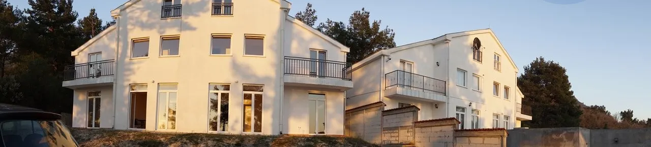 White three-story house with balconies in Kavac, Montenegro: modern building facade against a pine forest backdrop.
