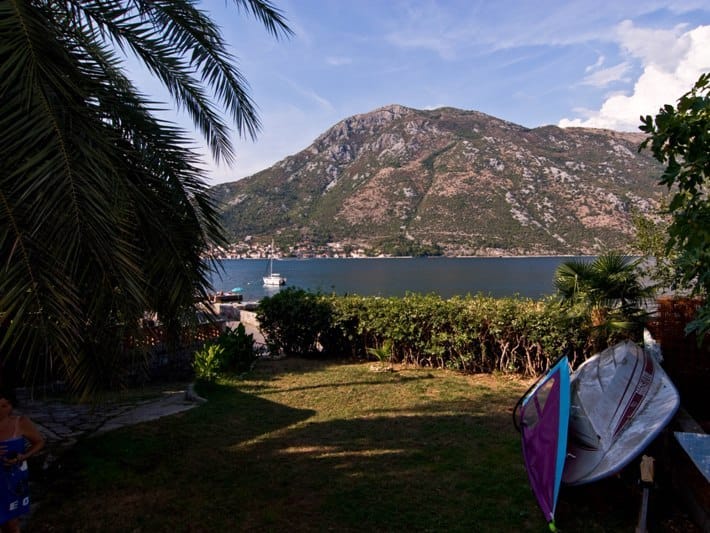 Pogled na Bokokotorski zaliv i planine iz bašte kuće u Stolivu, Crna Gora: palme, travnjak i čamac na obali mora.