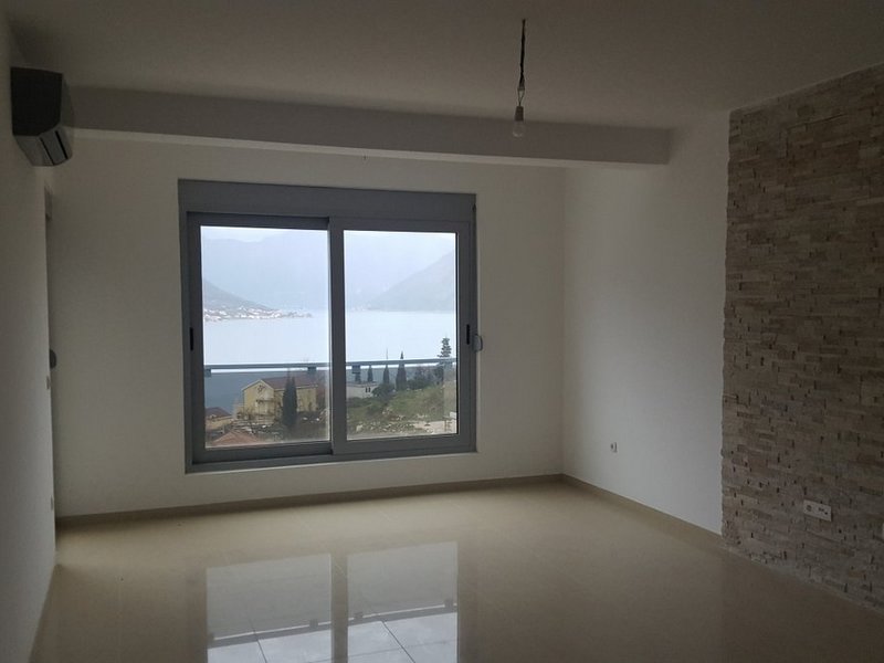 Wohnzimmer in einer Wohnung in Dobrota, Montenegro: helle Ausstattung, Steinwand, Panoramafenster mit Blick auf die Bucht von Kotor.