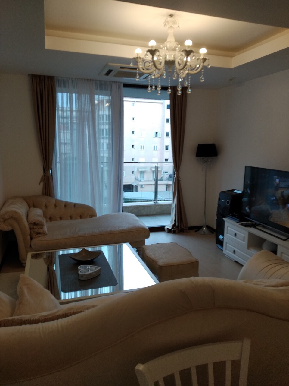 Dnevna soba sa bež nameštajem, staklenim stolom i lusterom u stanu u Budvi, Crna Gora, sa izlazom na balkon.