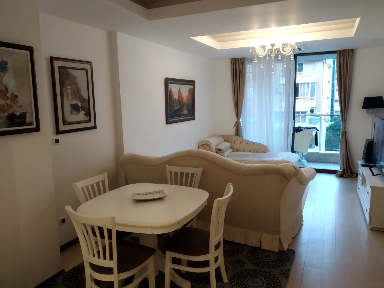 Dnevna soba sa trpezarijskim delom u stanu u Budvi, Crna Gora: svetao nameštaj, sofa, slike i izlaz na balkon.