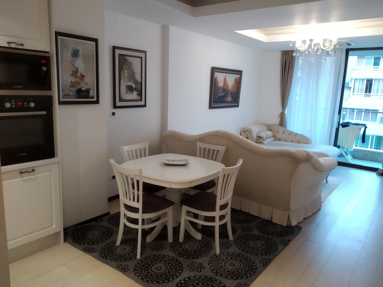 Svetao dnevni boravak sa trpezarijom i kuhinjom u stanu, Budva, Crna Gora: beli nameštaj, sofa i izlaz na balkon.