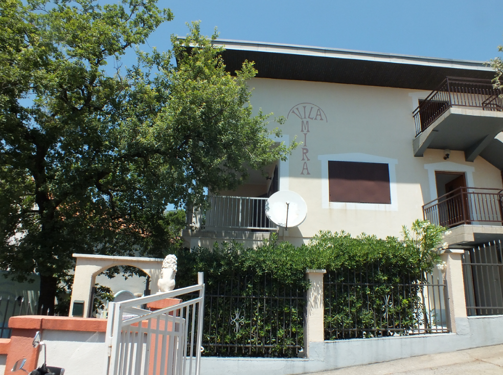 Façade de la villa à deux étages Vila Mira dans la ville de Bar, Monténégro, avec balcons, clôture en fer forgé et arbres verts.