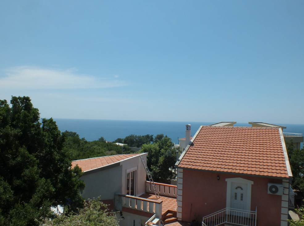 Vue sur la mer et la terrasse d’une maison au toit en tuiles à Bar, Monténégro, entourée de verdure et d’arbres.