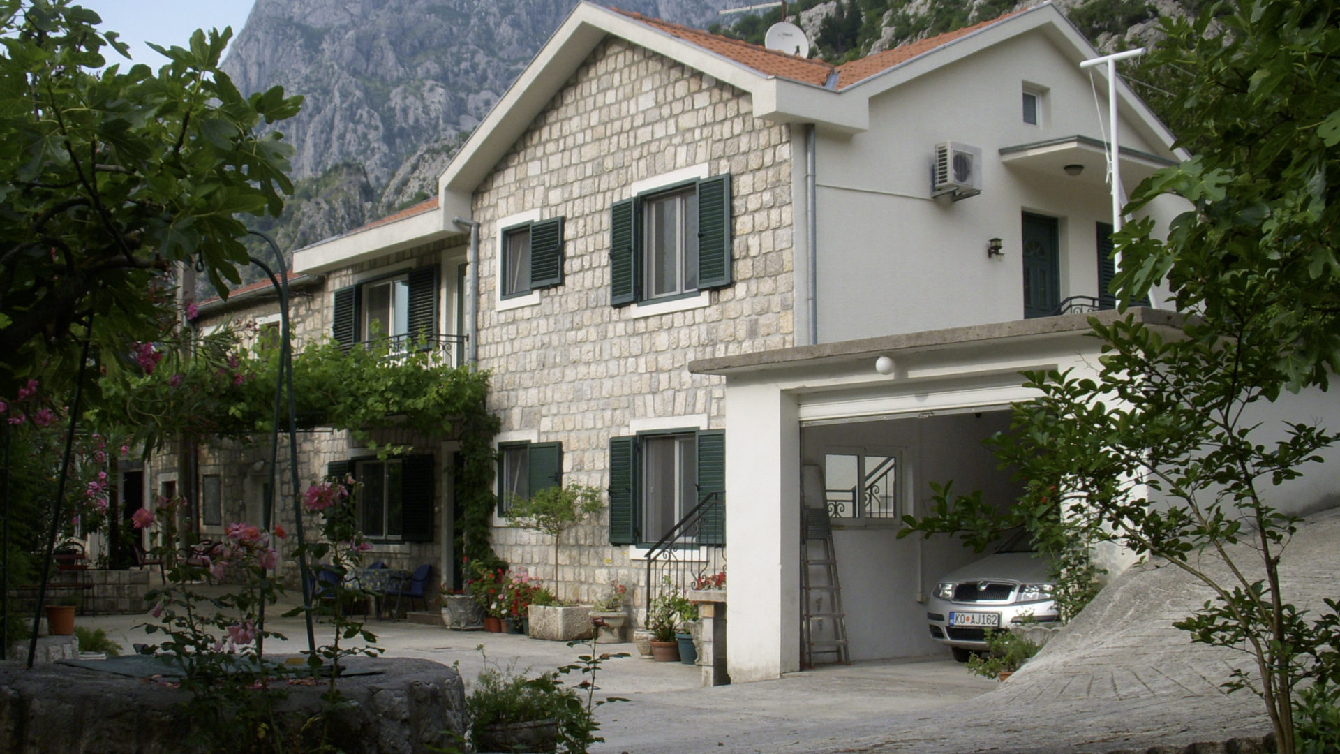 Zweistöckiges Steinhaus mit Garage und Garten in Kotor, Montenegro, vor Bergkulisse.
