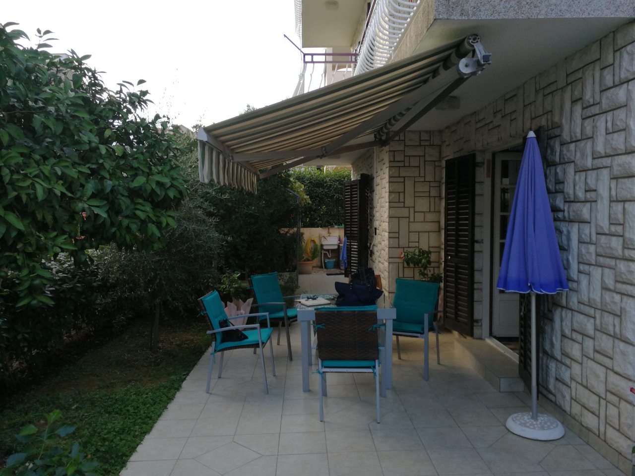Gemütliche Terrasse mit Gartenmöbeln und Überdachung am Haus in Tivat, Montenegro, mit einer mit Stein verkleideten Wand und grünem Garten.