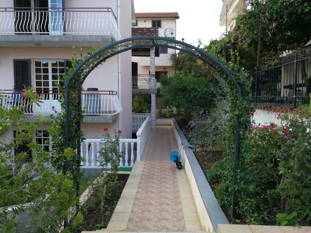 Grüne Gartenlaube und gepflasterter Weg im Hof eines Privathauses in Tivat, Montenegro, mit Blick auf die Fassade des Gebäudes.