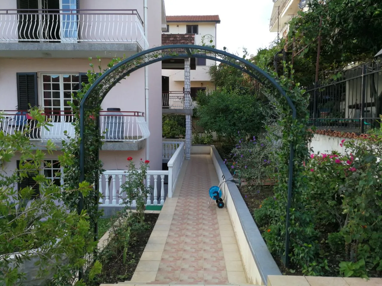 Grüne Gartenlaube und gepflasterter Weg im Hof eines Privathauses in Tivat, Montenegro, mit Blick auf die Fassade des Gebäudes.