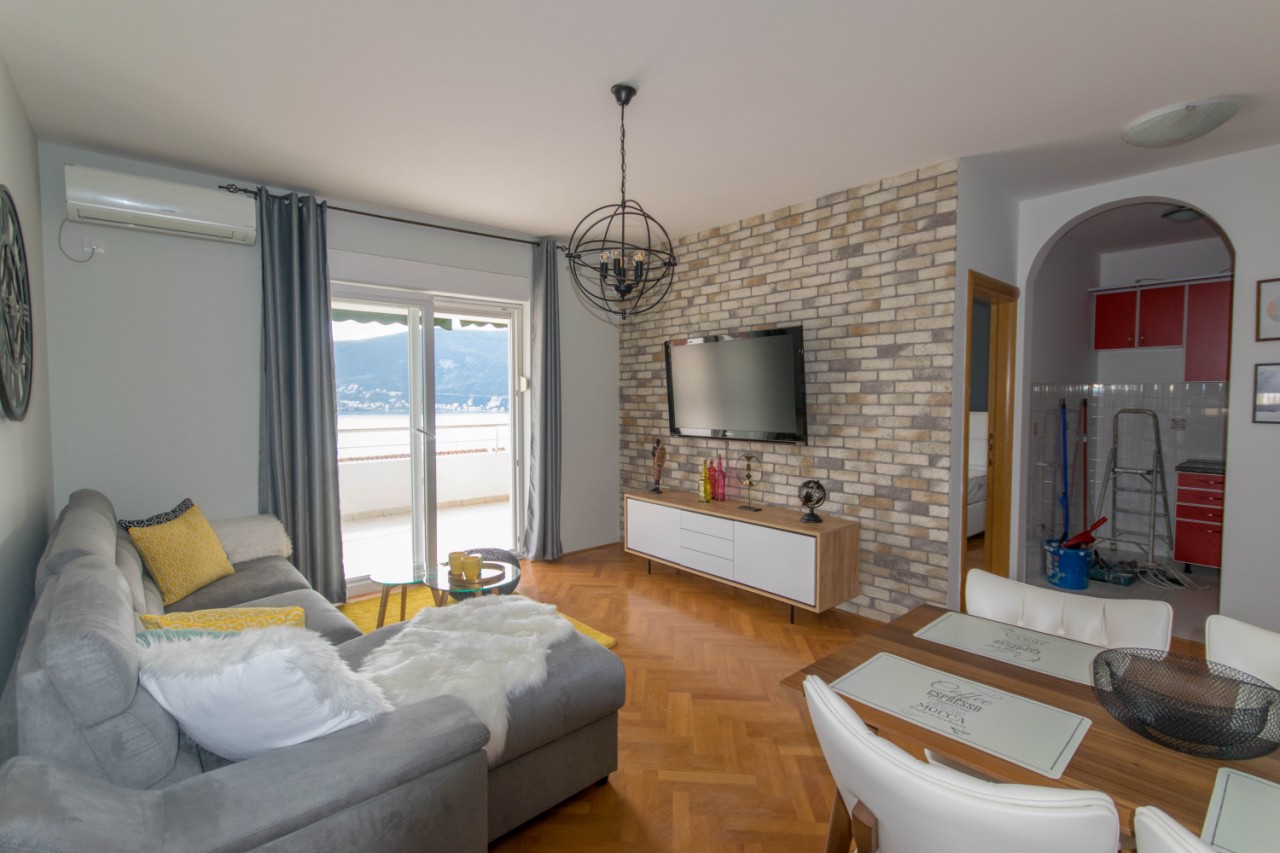 Wohnzimmer mit Meerblick in einer Wohnung in Herceg Novi, Montenegro: Ziegelwand, graues Sofa und Essbereich.