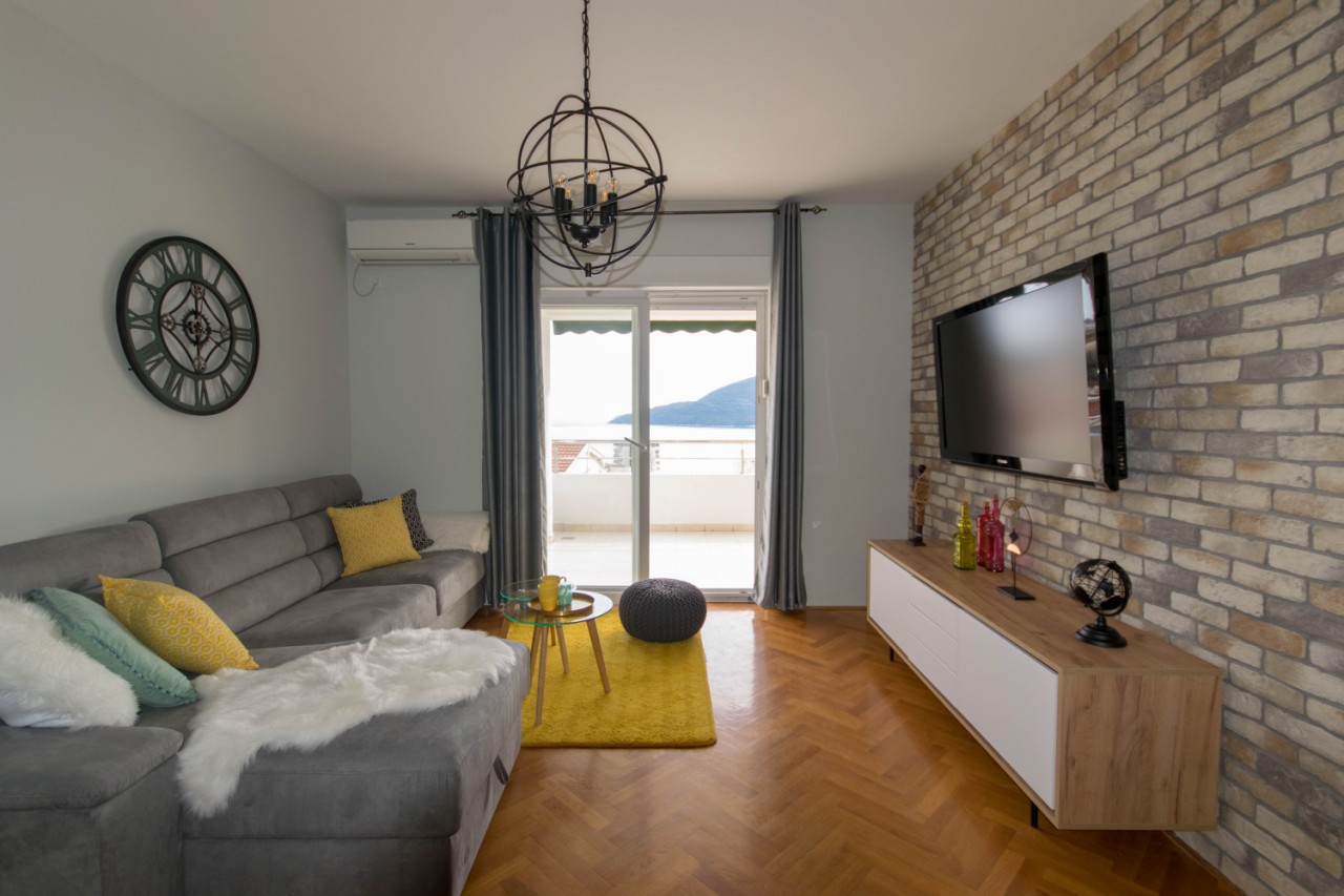 Modernes Wohnzimmer in einer Wohnung in Herceg Novi, Montenegro: Ziegelwand, graues Sofa und Meerblick aus dem Fenster.