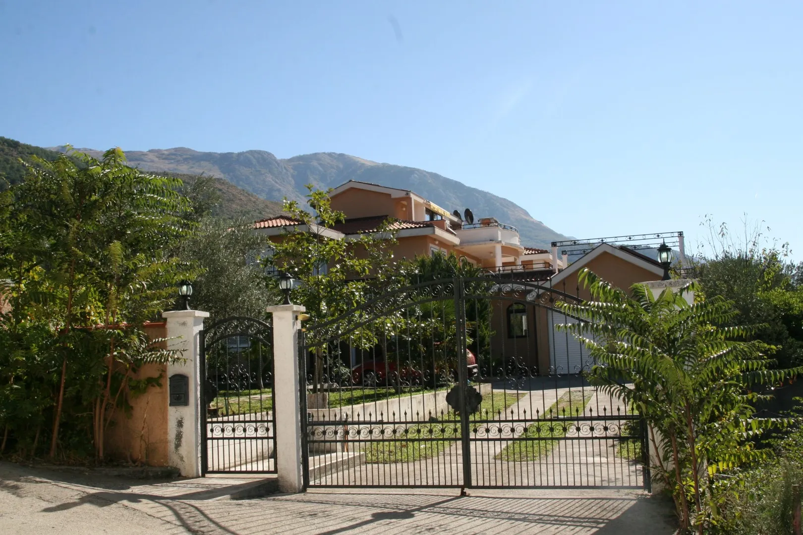 Ein großes Wohnhaus vor einer Bergkulisse, durch ein Metalltor gesehen.