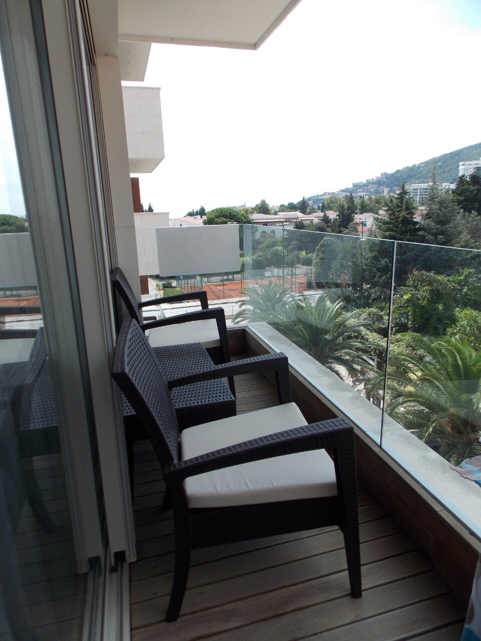 Balkon sa pletenim nameštajem u stanu u Budvi, Crna Gora, s pogledom na zeleni park i teniske terene.