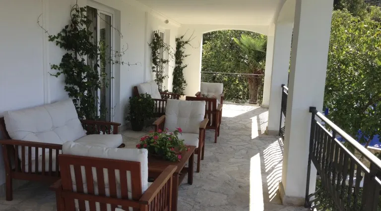 Ugodna terasa sa drvenim nameštajem i pogledom na baštu u vili u Bečićima, Crna Gora. Kamena obloga poda i zelenilo.