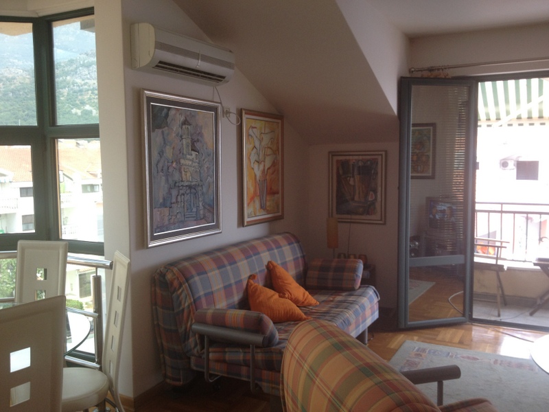 Wohnzimmer in einer Wohnung in Budva, Montenegro: karierter Sofa, Bilder an den Wänden, Klimaanlage und Zugang zum Balkon.