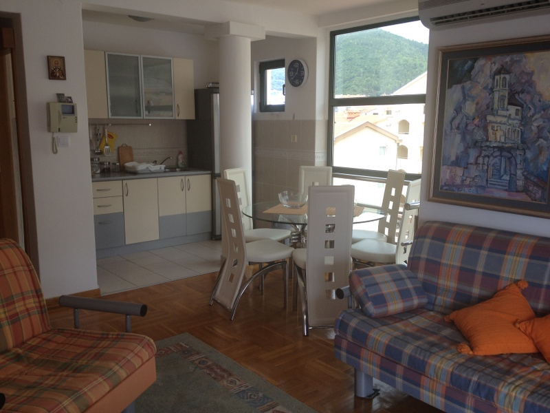 Wohnzimmer mit Küche in einer Wohnung in Budva, Montenegro: helle Möbel, Essbereich und Bergblick durch das Panoramafenster.