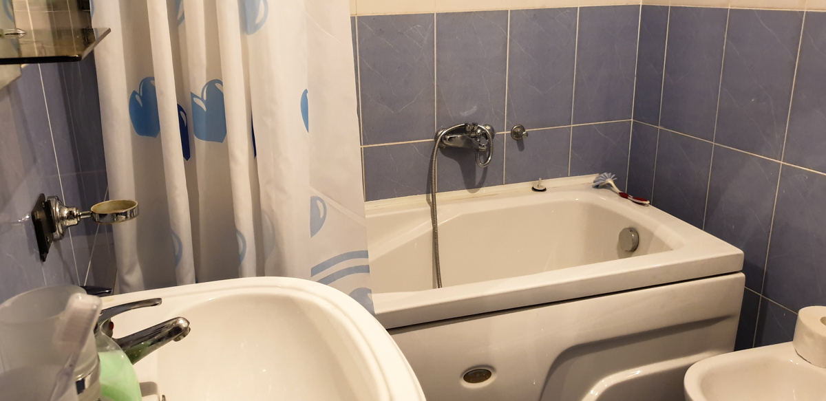 Kupatilo u stanu u gradu Bar, Crna Gora: bela kada, plave pločice na zidovima i zavesa sa printom.