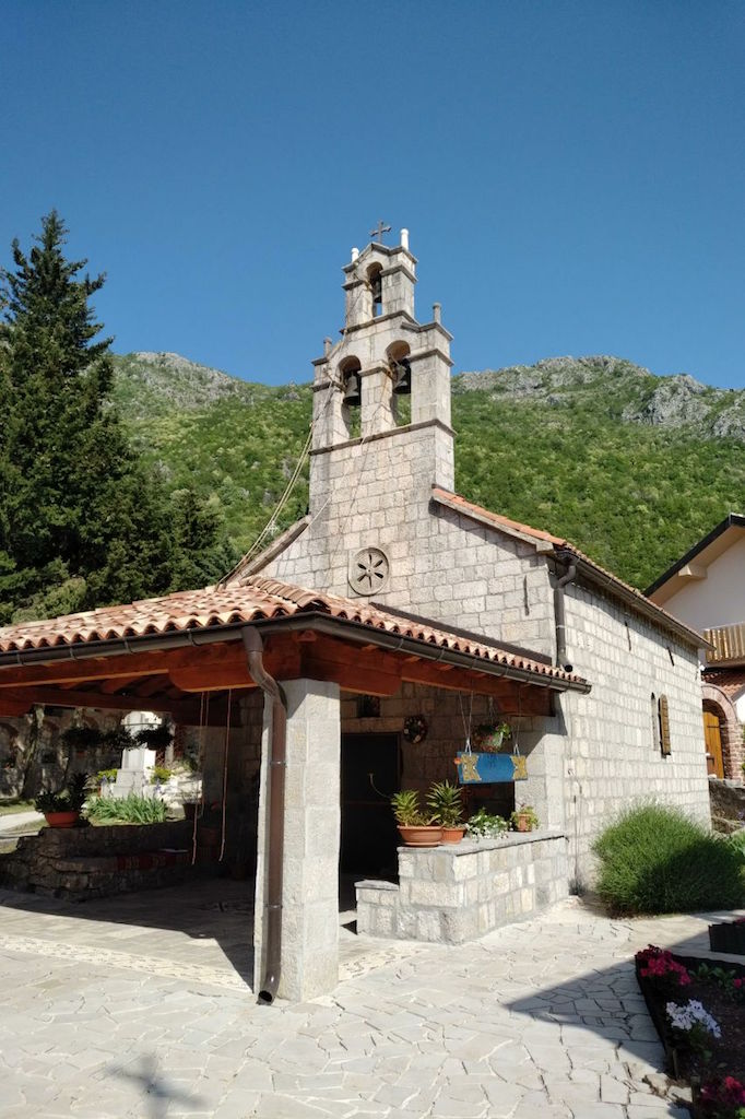 Stara kamena crkva pored placa u Svetom Stefanu, Crna Gora, u pozadini zelenih planina i vedrog neba.