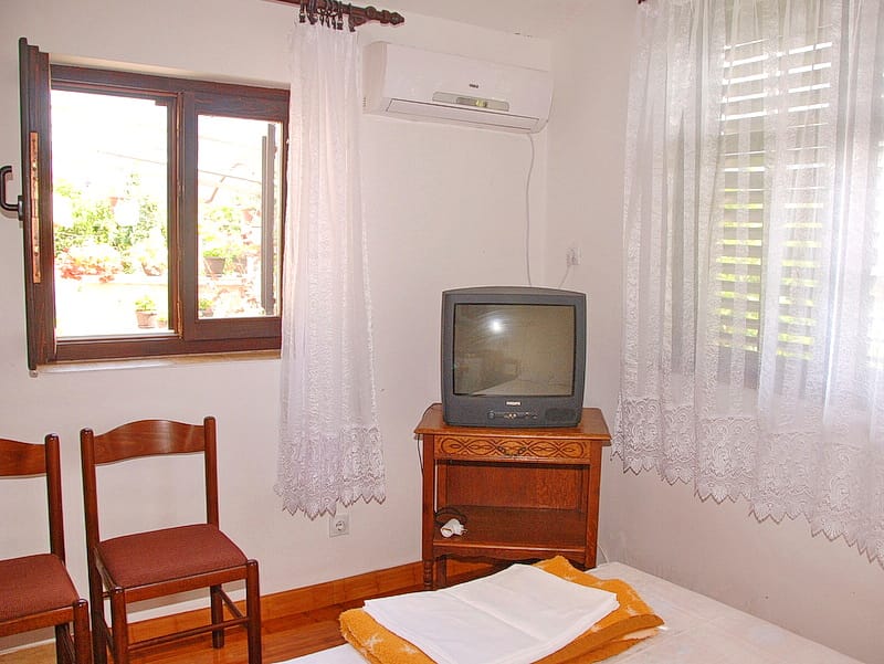Chambre dans une maison à Igalo, Monténégro : chaises en bois, meuble TV, climatisation et vue sur la terrasse avec des plantes.