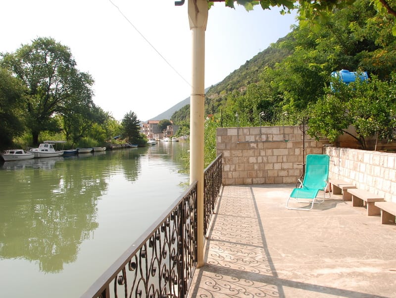 Terrasse d'une maison à Igalo, Monténégro : vue sur la rivière avec des bateaux, maçonnerie en pierre, chaise longue et garde-corps en fer forgé.