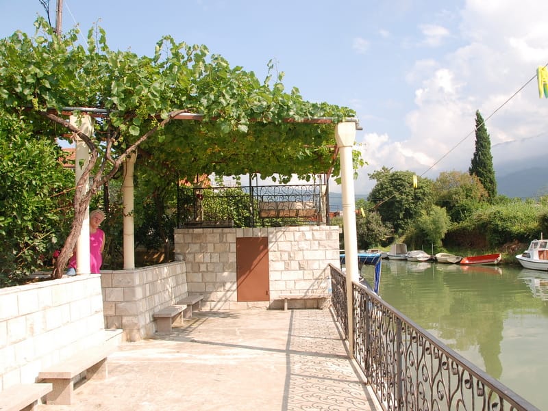 Terrasse avec vigne et bancs en pierre au bord de la rivière à Igalo, Monténégro, vue sur le quai avec des bateaux.