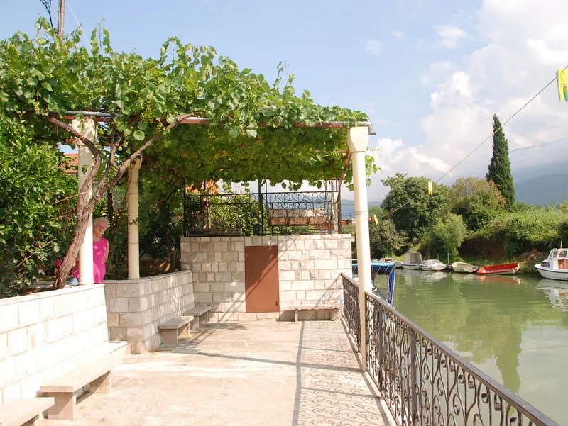Terrasse avec vigne et bancs en pierre au bord de la rivière à Igalo, Monténégro, vue sur le quai avec des bateaux.