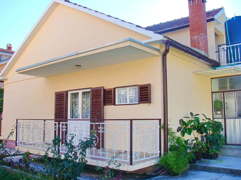 Single-story house in Baošići, Montenegro: light facade, terrace with wrought-iron railing, and brown shutters on the windows.