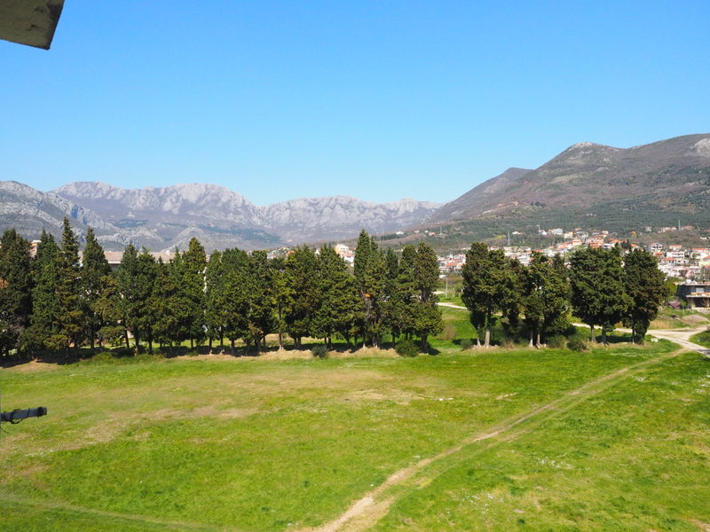 Panoramski pogled iz prozora stana u gradu Baru, Crna Gora: zelena livada, aleja čempresa i planinski pejzaž u pozadini.