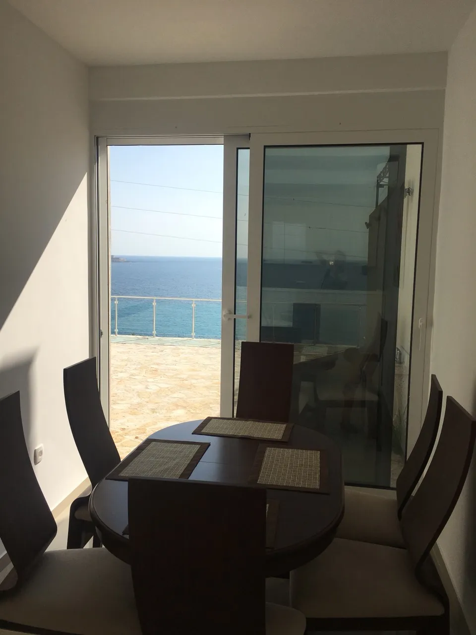 Dining area with a round table and chairs next to a glass door opening to a balcony overlooking the sea.