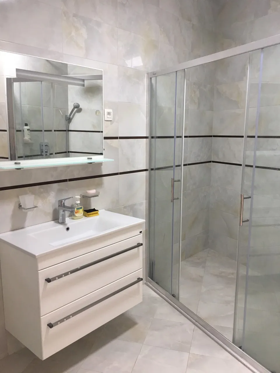 Bathroom featuring a white vanity sink, mirror with shelf, and a glass shower enclosure against tiled walls.