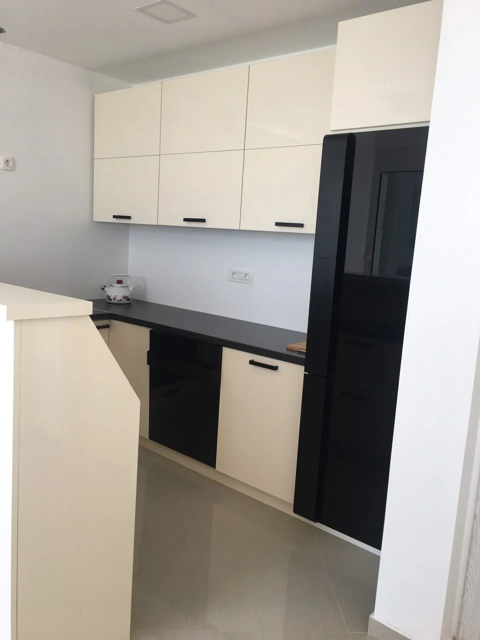 Modern kitchen with beige glossy cabinets, black countertop, built-in black refrigerator, and a white teapot.