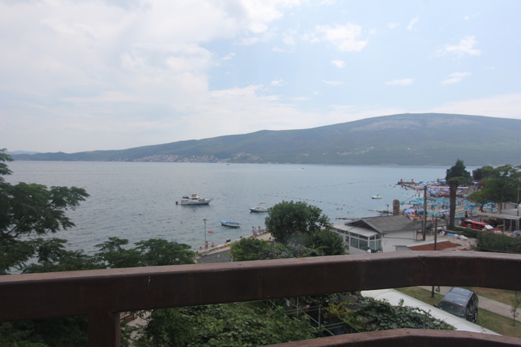 Blick vom Balkon eines Mini-Hotels in Baošići, Montenegro: Panorama von Meer, Strand und Bergen, Holzgeländer im Vordergrund.