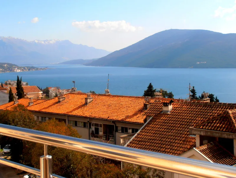 Pogled sa balkona stana u Herceg Novom, Crna Gora: panorama Bokokotorskog zaliva, planina i krovova kuća od crepova.