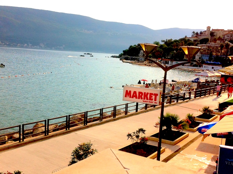 Blick auf die Uferpromenade, den Strand und das Meer in Herceg Novi, Montenegro: Flanierzone mit Palmen und einem Geschäft an der Küste.