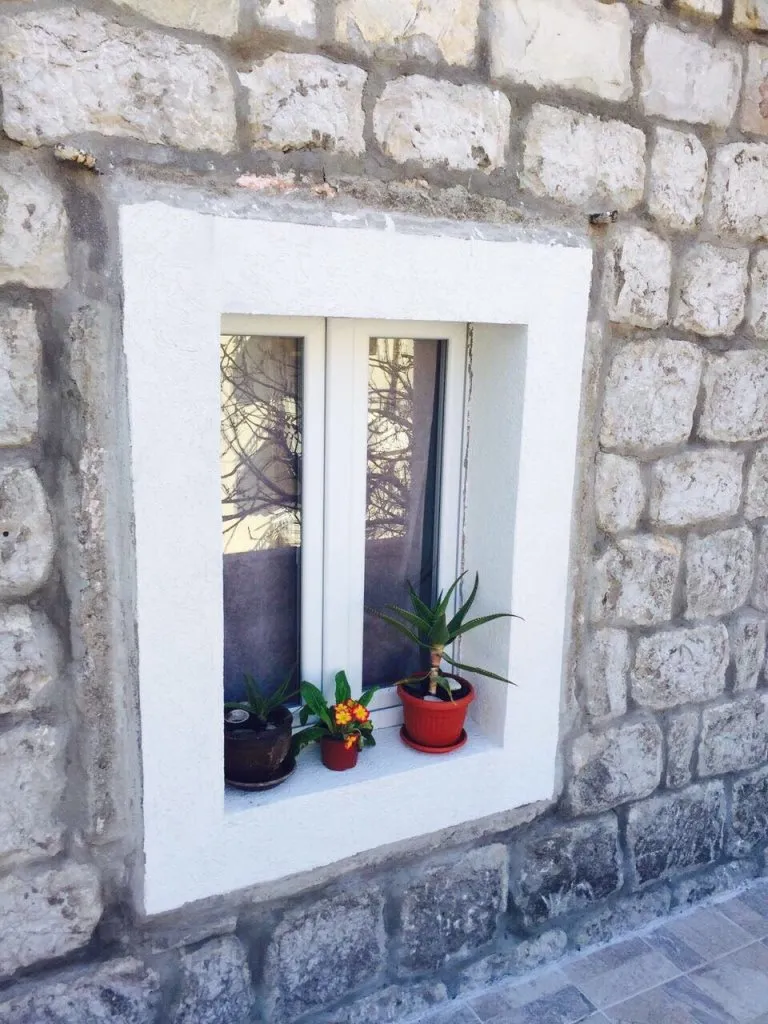 Fenêtre en plastique blanche dans un mur en pierre d’un mini-hôtel à Rafailovici, Monténégro, avec des plantes d’intérieur sur le rebord de la fenêtre.