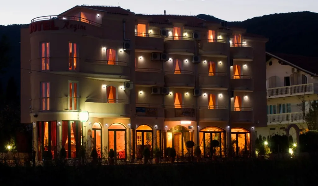 Fassade des Hotels Regina in Bijela, Montenegro: abendliche Beleuchtung des Gebäudes, Balkone und Panoramafenster vor Bergkulisse.