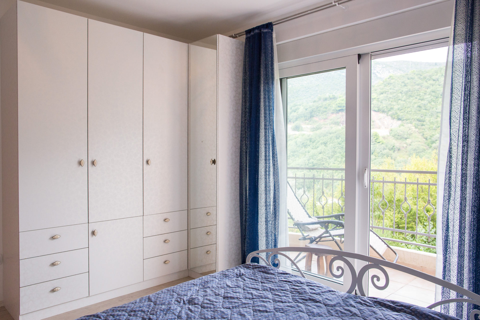 Spavaća soba u stanu u Bečićima, Crna Gora: beli ormar, kovani krevet i izlaz na balkon s pogledom na planine.