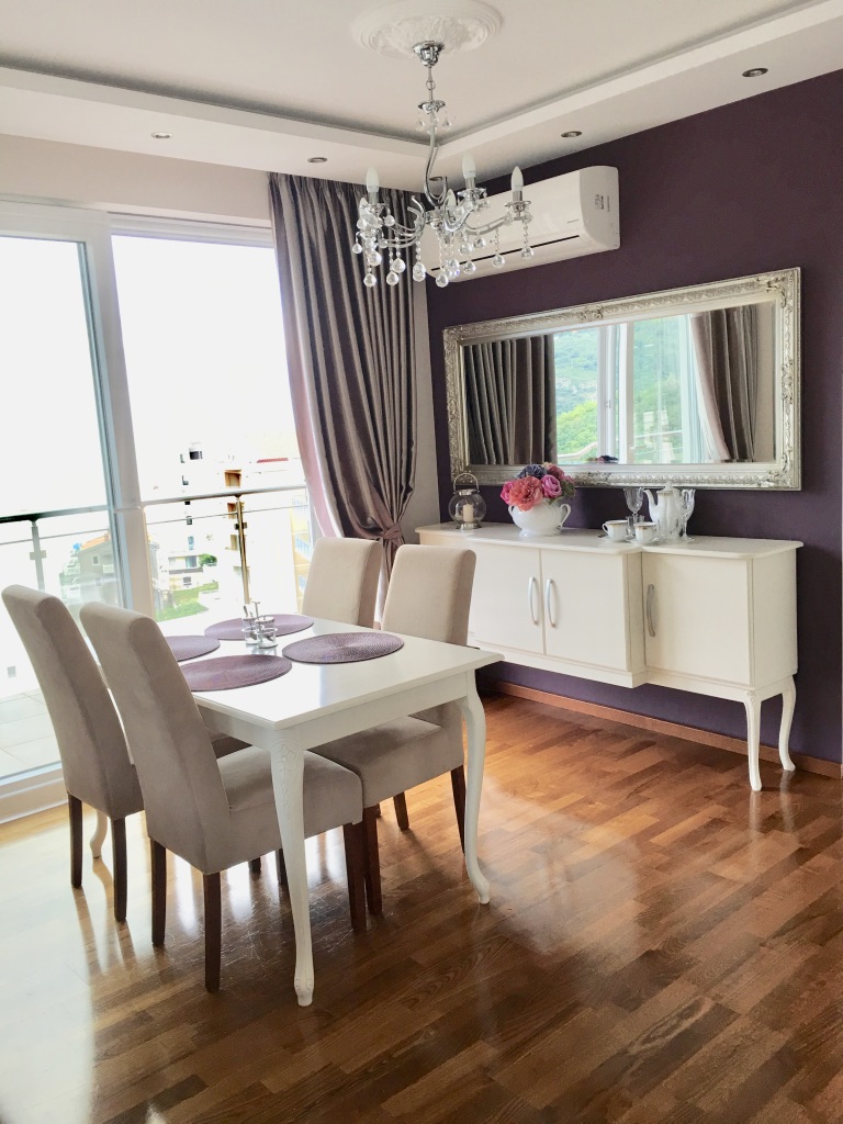 Heller Essbereich in einer Wohnung in Bečići, Montenegro: weiße Möbel, Parkett, violette Wand und Panoramafenster.